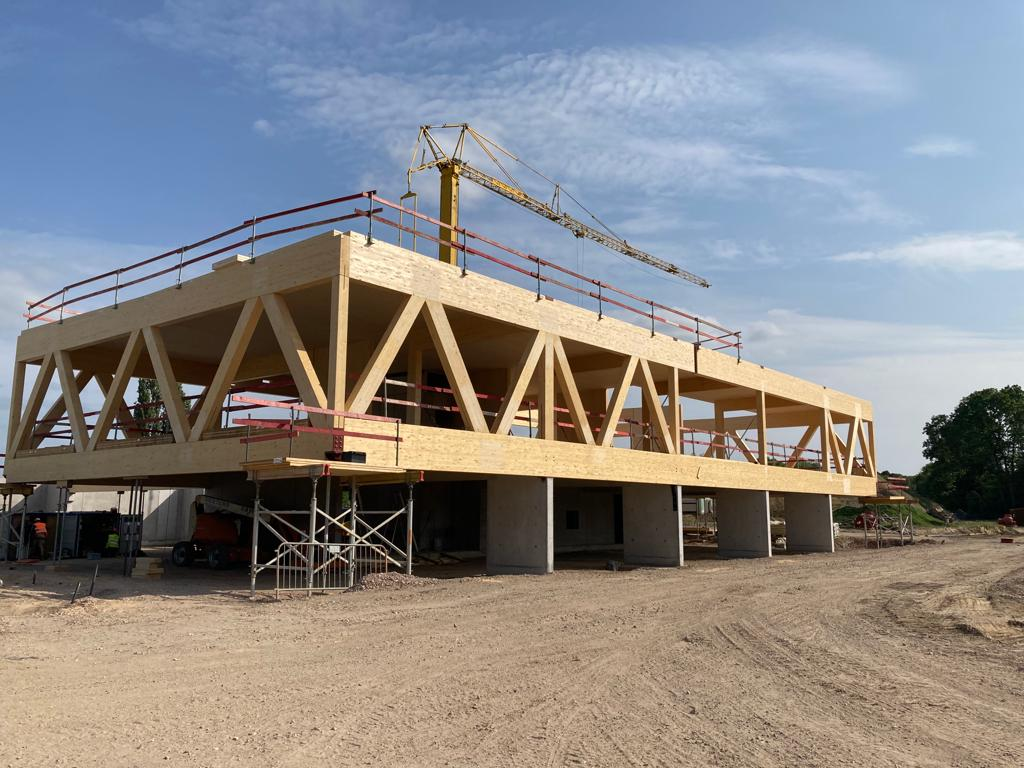 Houten draagstructuur met vakwerkliggers op een werf, met torenkraan op de achtergrond