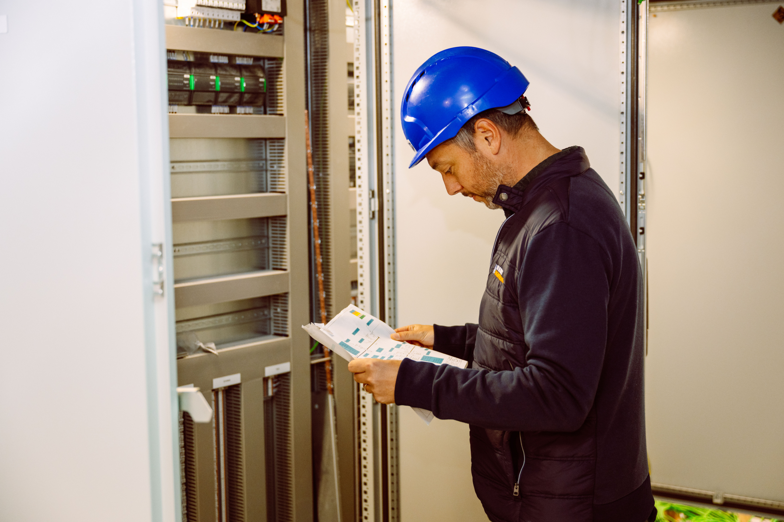 Ingenieur met blauwe helm leest een plan bij een open elektriciteitskast